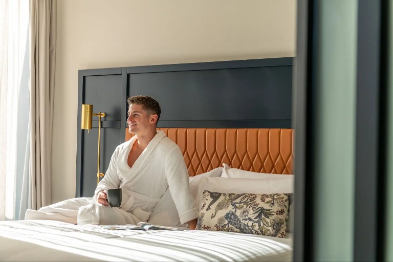 Man wearing a bathrobe holding a cup on Dorsett Hotel room bed.