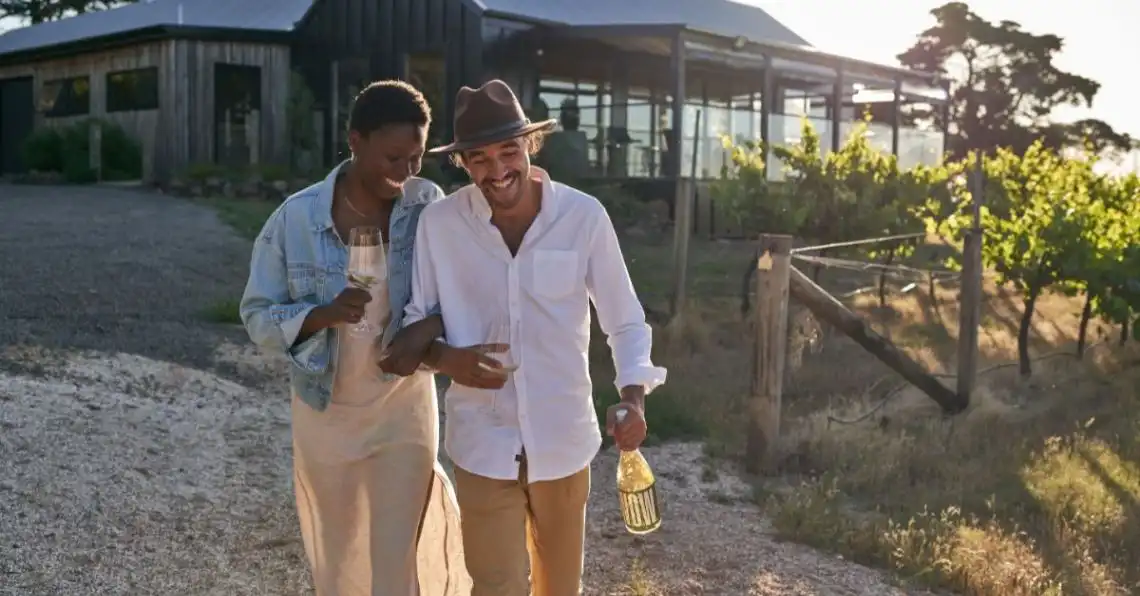 Couple laugh lovingly carrying a bottle and glass of wine through a winery