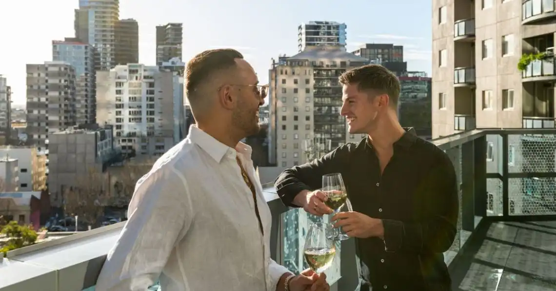 Two men sip golden wine on a Dorsett Melbourne balcony in the afternoon light