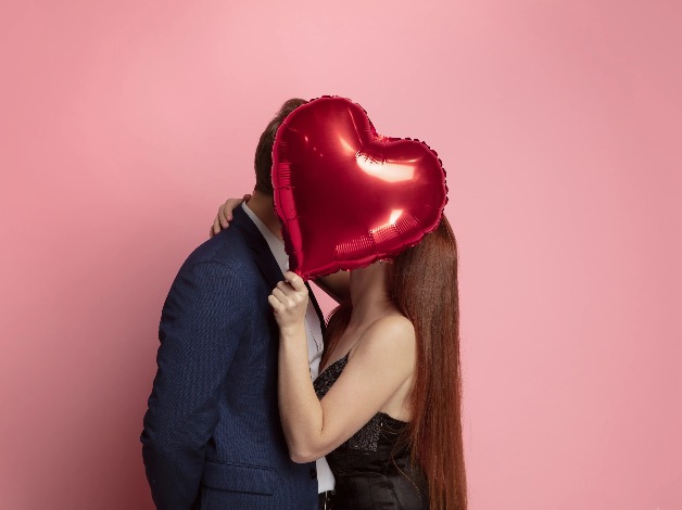 A couple hiding their faced behind a red heart balloon